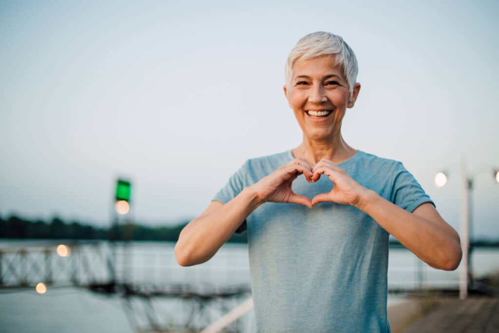 Smiling person with short gray hair making a heart shape with hands, standing near water with a blurred background of trees and a bridge.