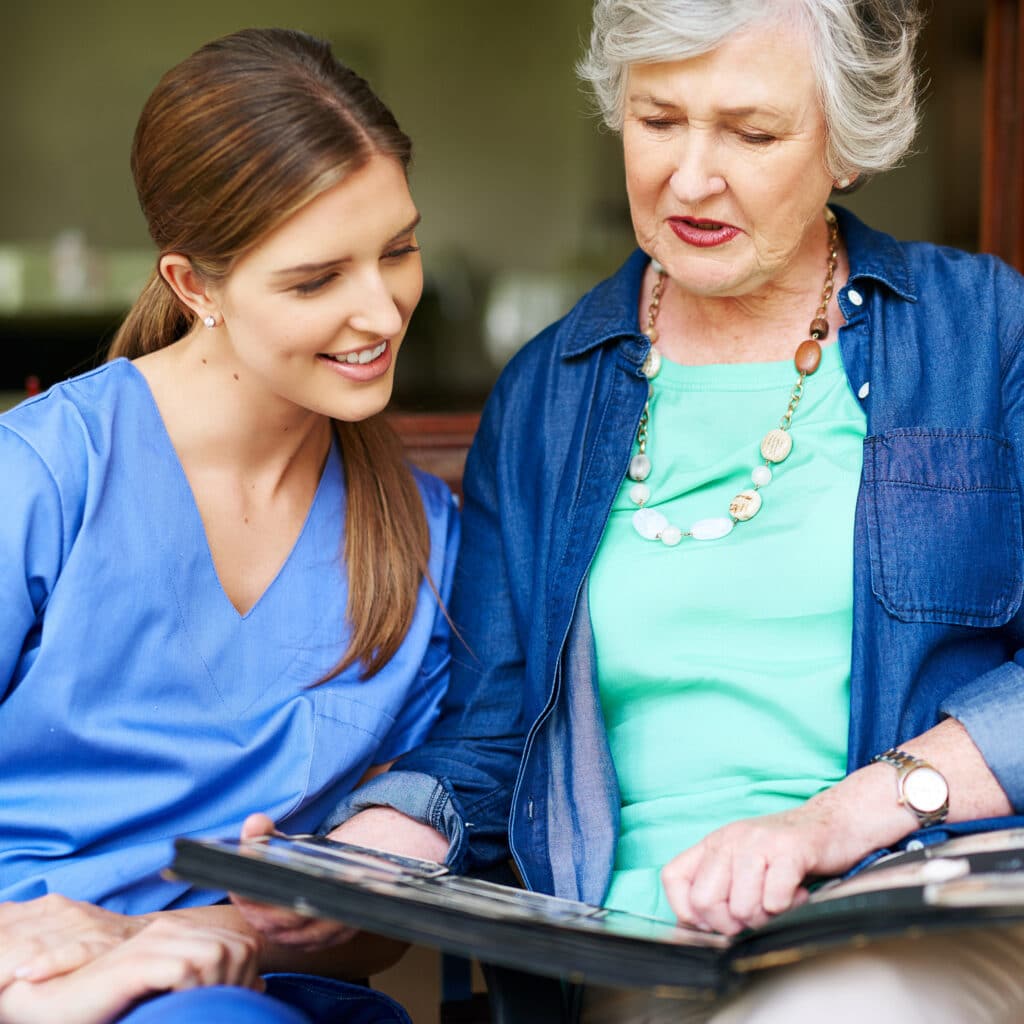 A young woman in blue scrubs and an older woman in a denim shirt are sitting together, looking at a photo album and smiling.