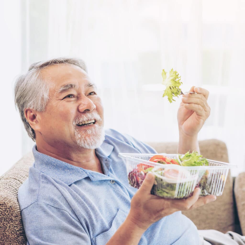 Older man smiling and holding a fork with lettuce, sitting on a couch with a salad bowl.