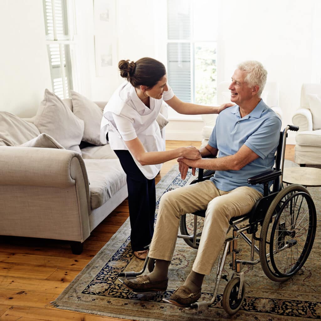 A caregiver interacts with an elderly man in a wheelchair in a living room setting.