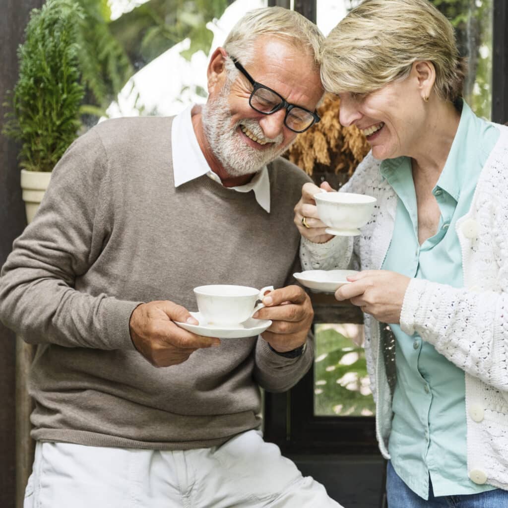 Older couple smiling and holding teacups, standing near a window with a plant nearby.