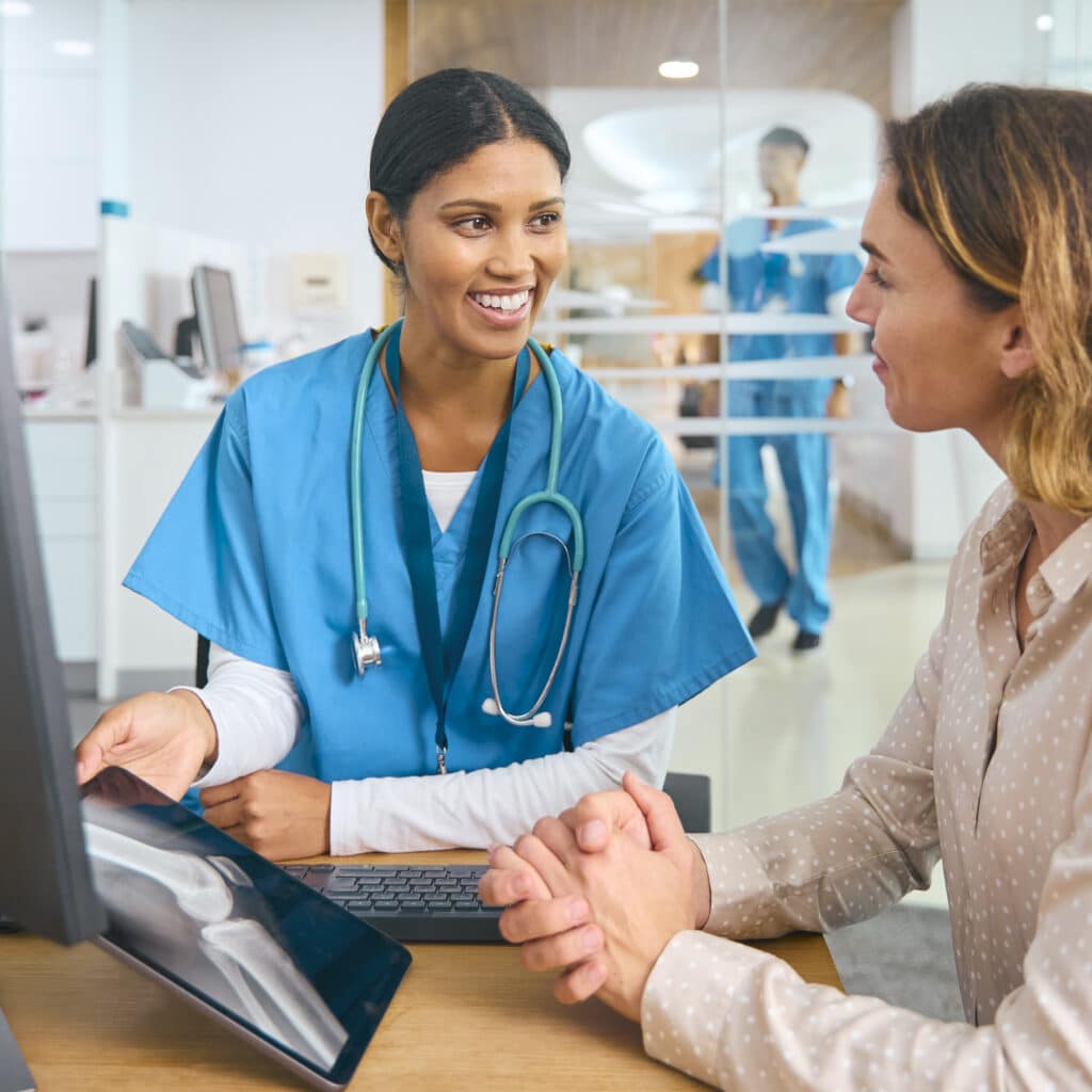 A healthcare professional shows an X-ray to a patient at a desk. Both are engaged in conversation. Another medical professional is visible in the background.