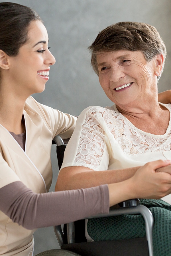 A young woman and an older woman in a wheelchair share a smile indoors, embodying the warmth of quality care.